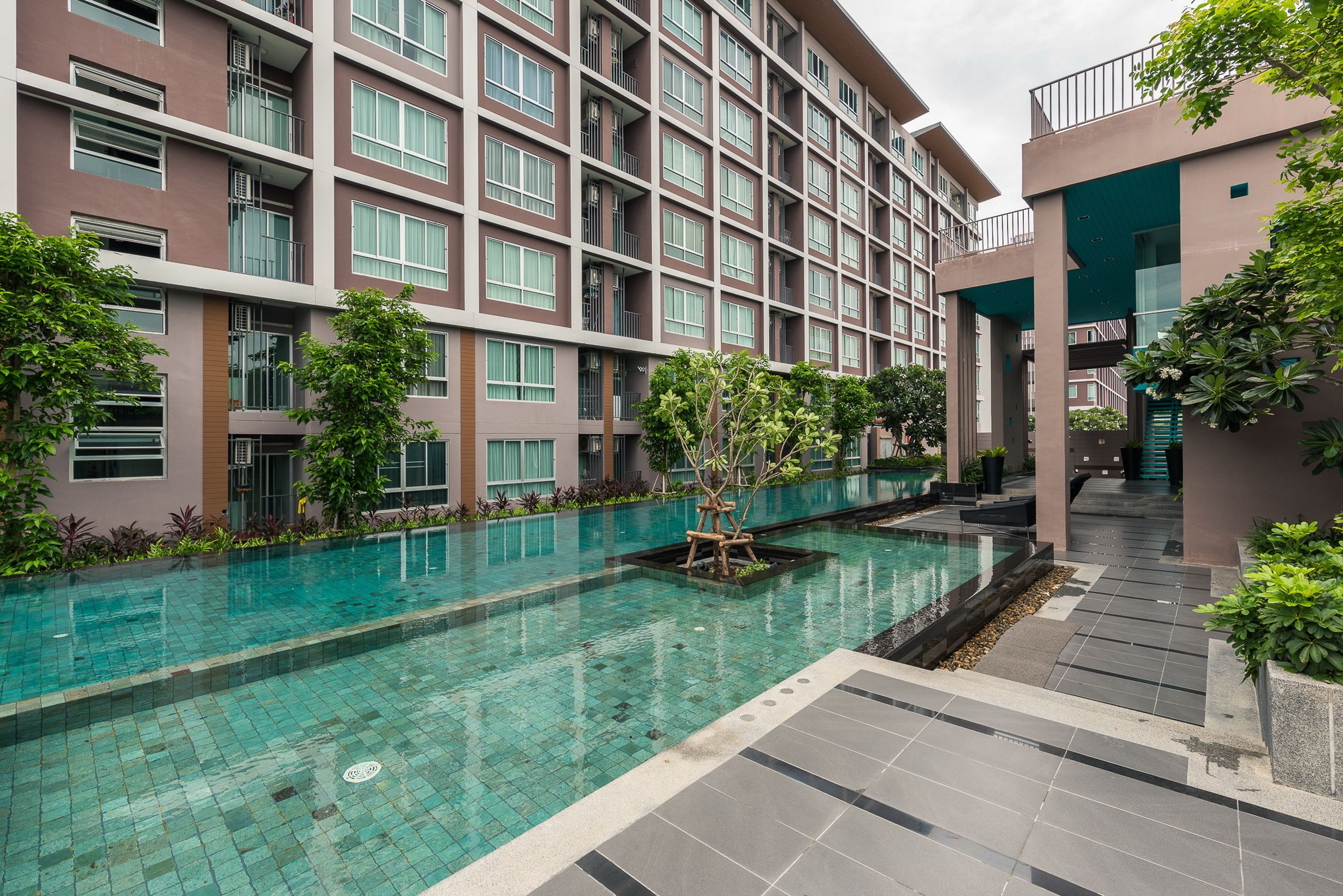 A modern hotel with a swimming pool, surrounded by greenery and a multi-story building with balconies.