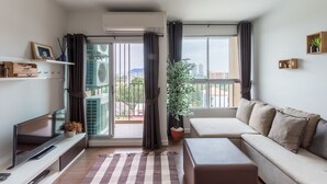 A modern living room with a sofa, a flat-screen TV, a potted plant, and a balcony with a view of the city.