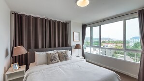 A hotel room with a large bed, two bedside lamps, a nightstand, and a view of the city through large windows.