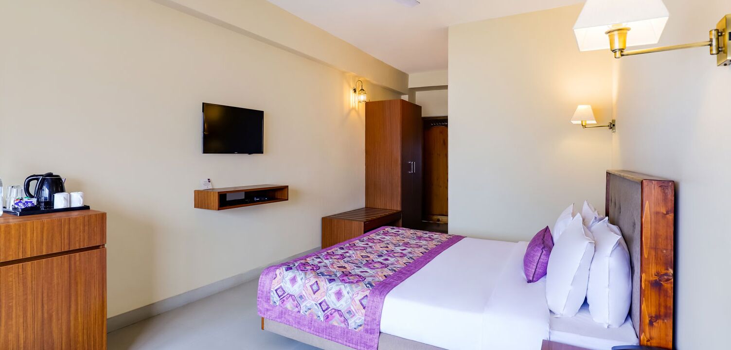 Een hotelkamer met een bed, een aan de muur bevestigde televisie, een houten garderobe, een nachtkastje en een lamp.