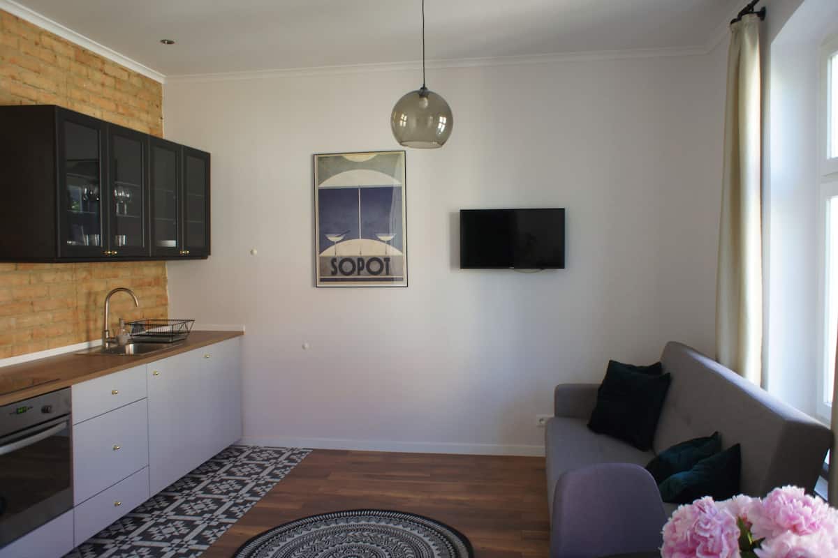 A modern kitchen with a brick backsplash, a dining area with a patterned rug, a grey sofa with dark cushions, and a flat-screen TV mounted on the wall.