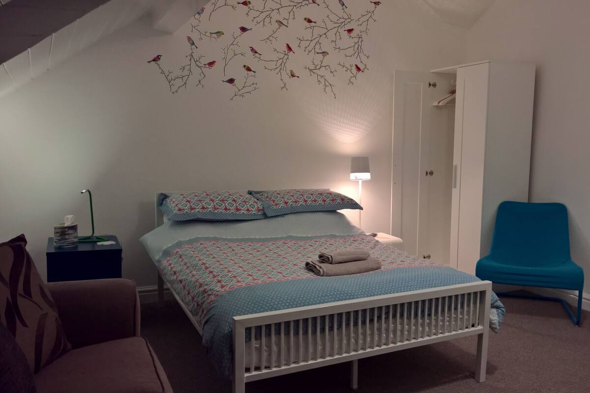 A bedroom with a bed, a chair, a bedside table, a lamp, and a wall decoration.