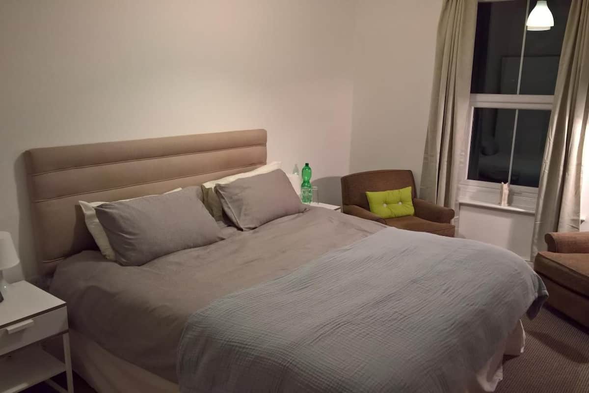 A neatly made bed with a headboard, a bedside table with a lamp, a chair with a pillow, and a window with curtains.