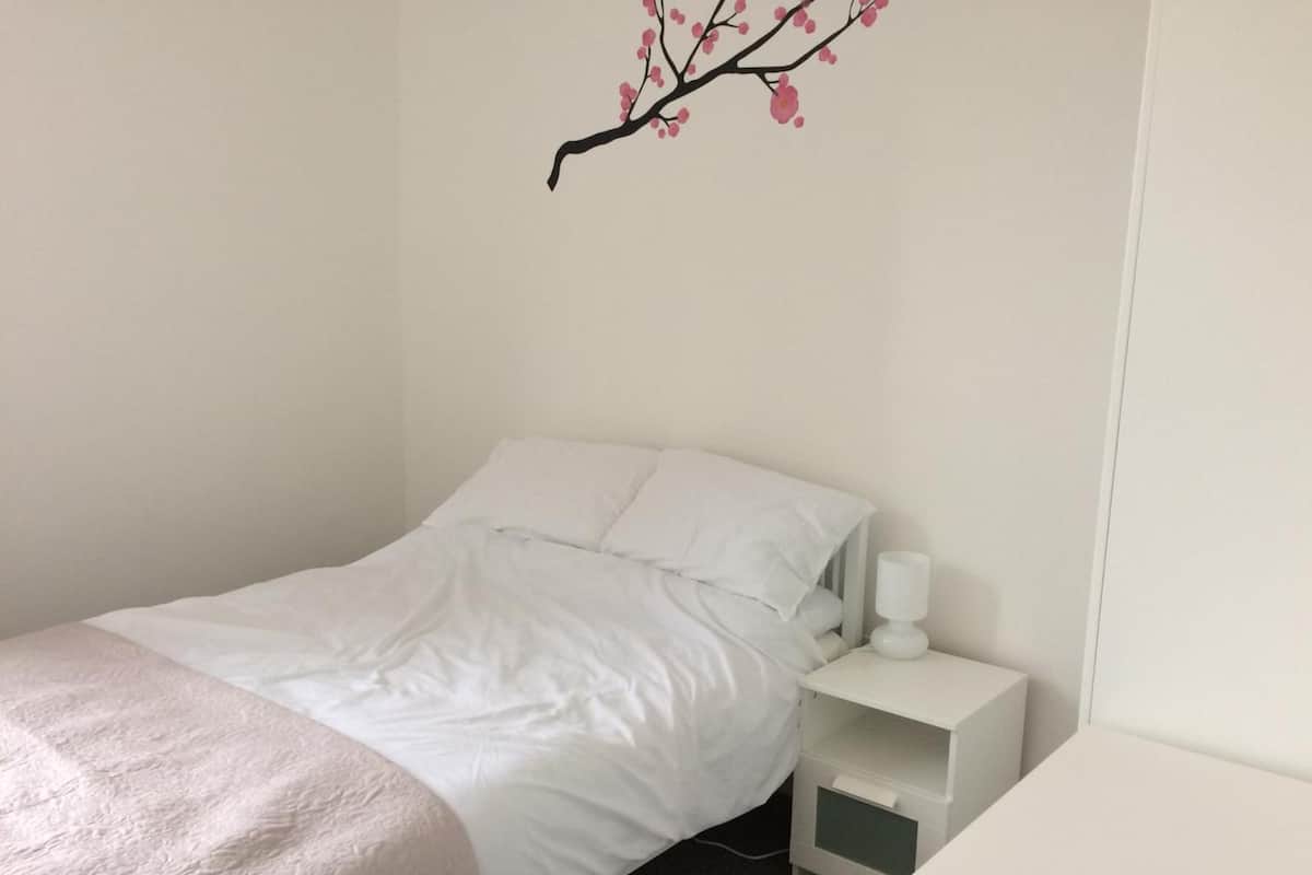 A neatly made bed with white bedding and a pink throw blanket, a white nightstand with a lamp, and a wall-mounted branch decoration.