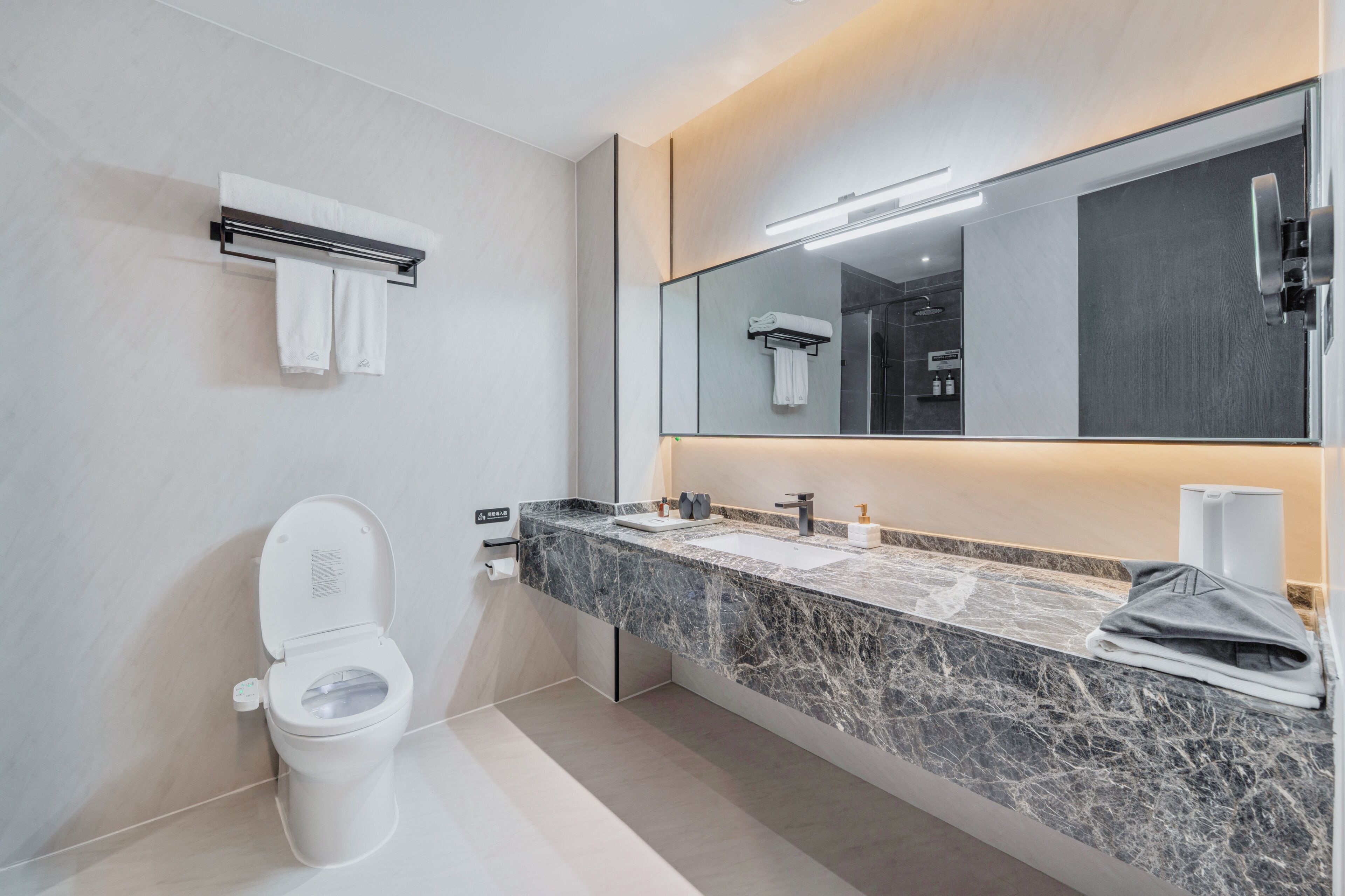Un baño moderno con inodoro, lavamanos con encimera de mármol, un espejo grande y un perchero para toallas.