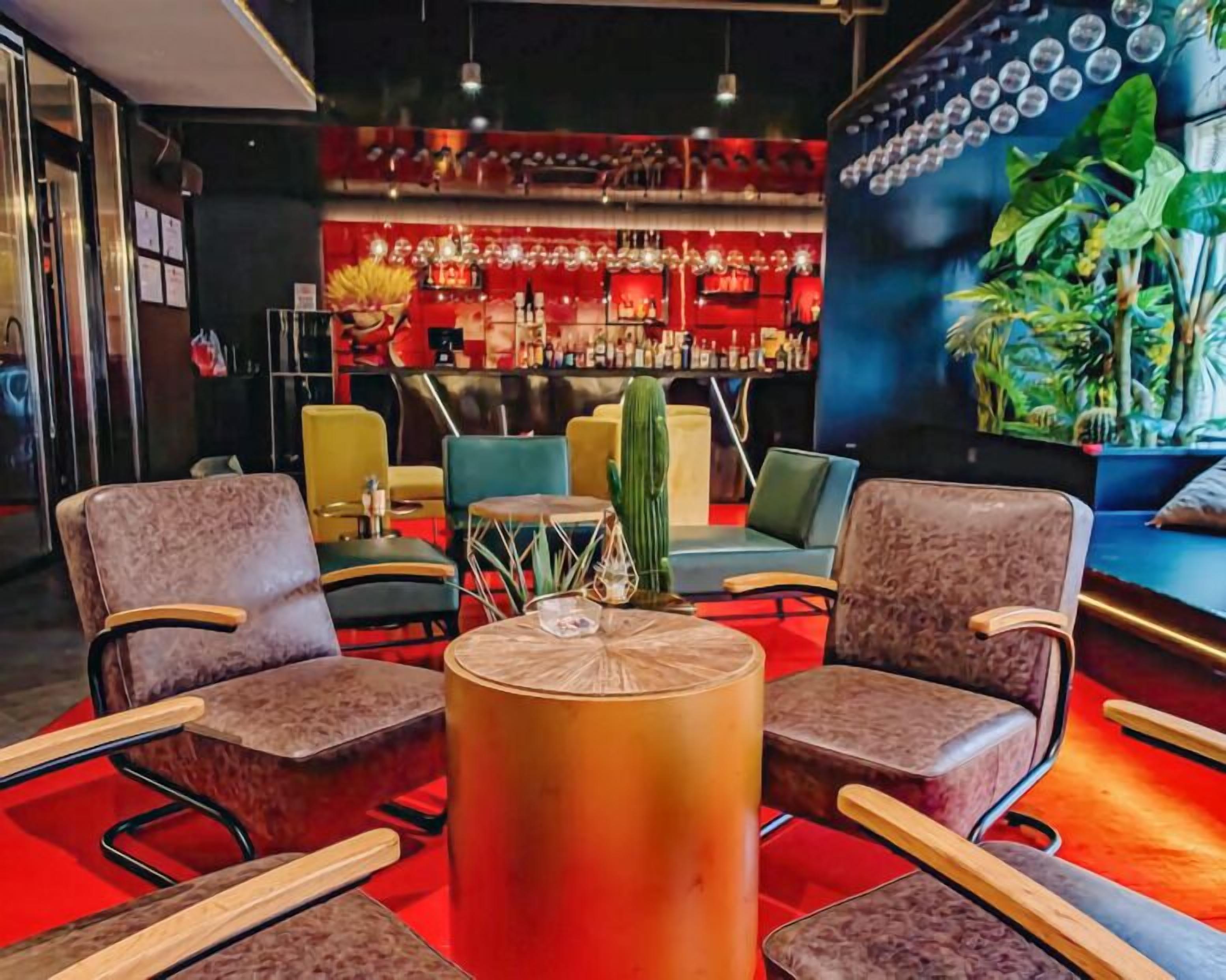 Zona de bar con asientos rojos y azules, una mesa redonda central y un fondo de luces colgantes y un mural de plantas.