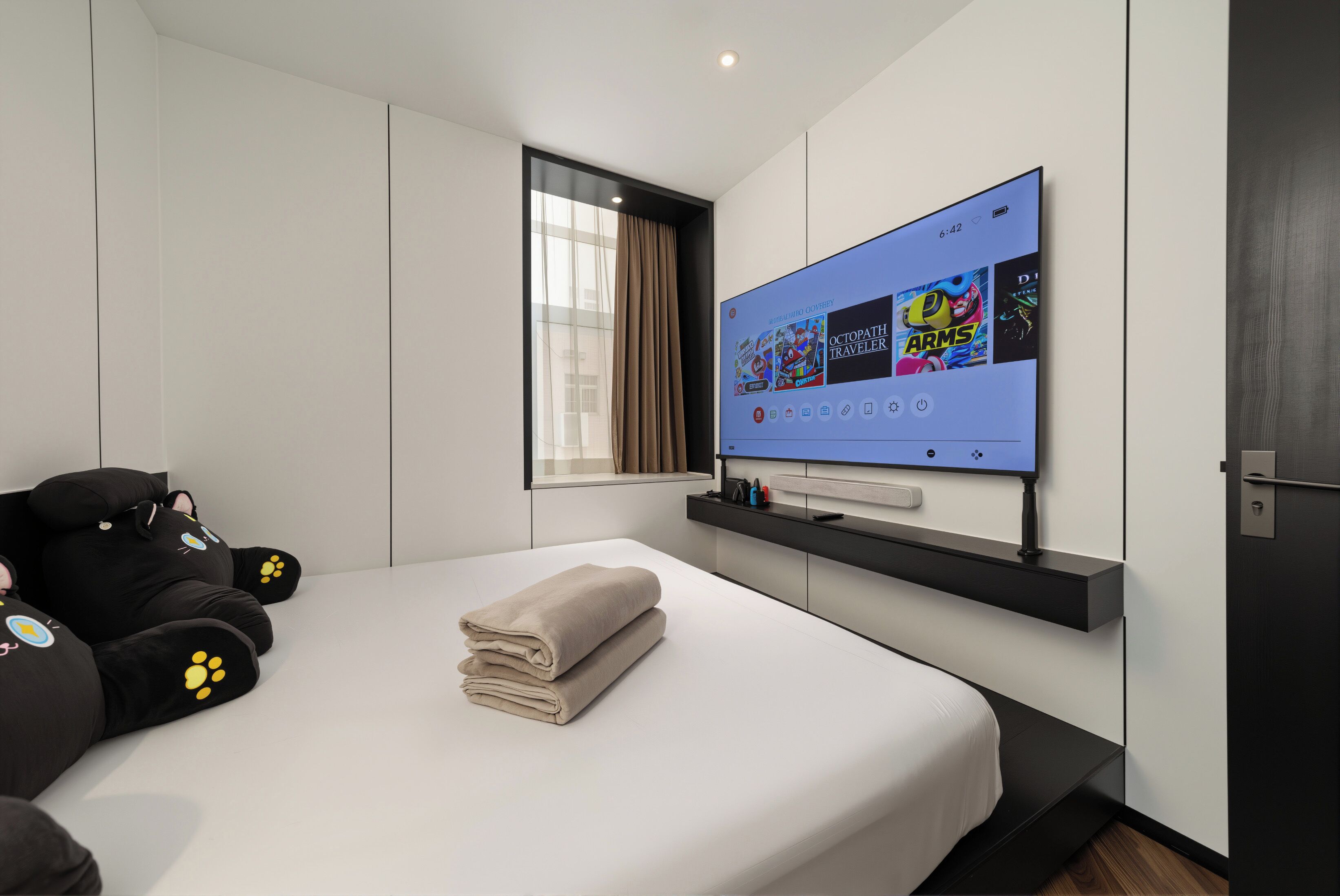A modern hotel room with a large bed, a flat-screen TV displaying game titles, a window with curtains, and a black sofa with paw prints.