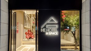 Der Eingang des HE HOTEL mit modernem Design, inklusive einer Neonreklame und einer Glastür.
