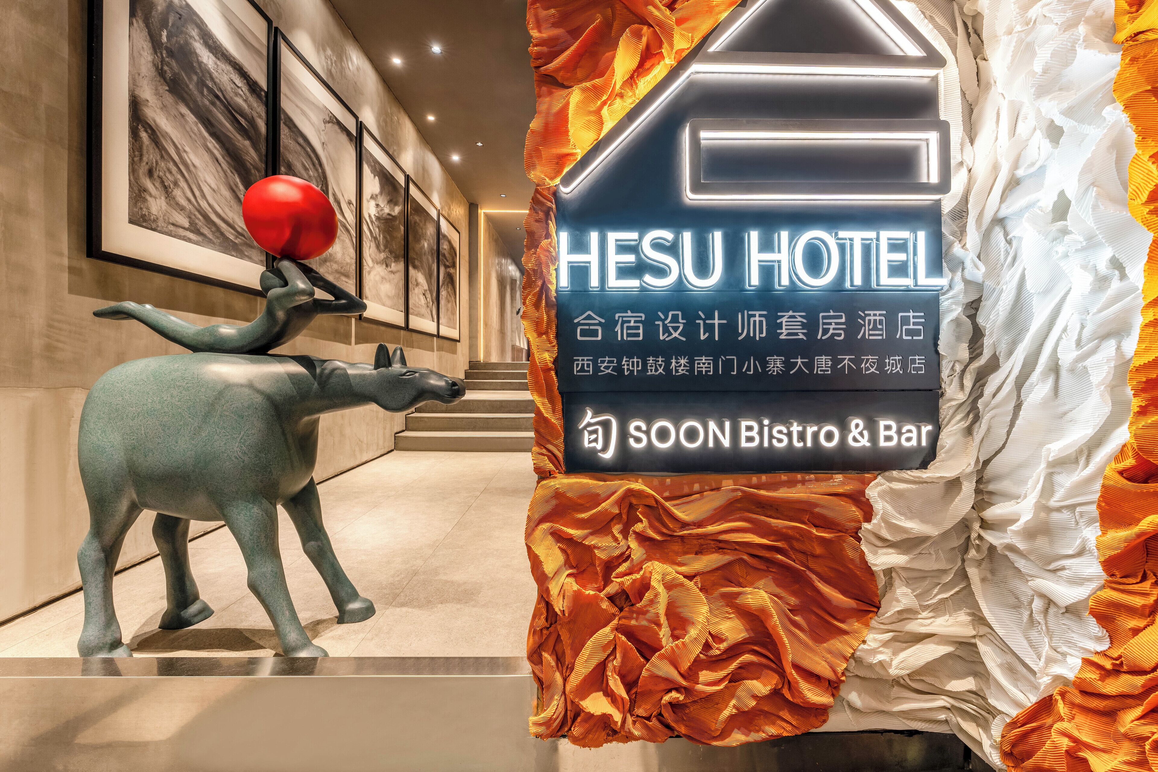 A hotel interior with a sculpture of a figure balancing a red ball on its head, framed artwork on the walls, and a sign indicating a soon-to-open bistro and bar.