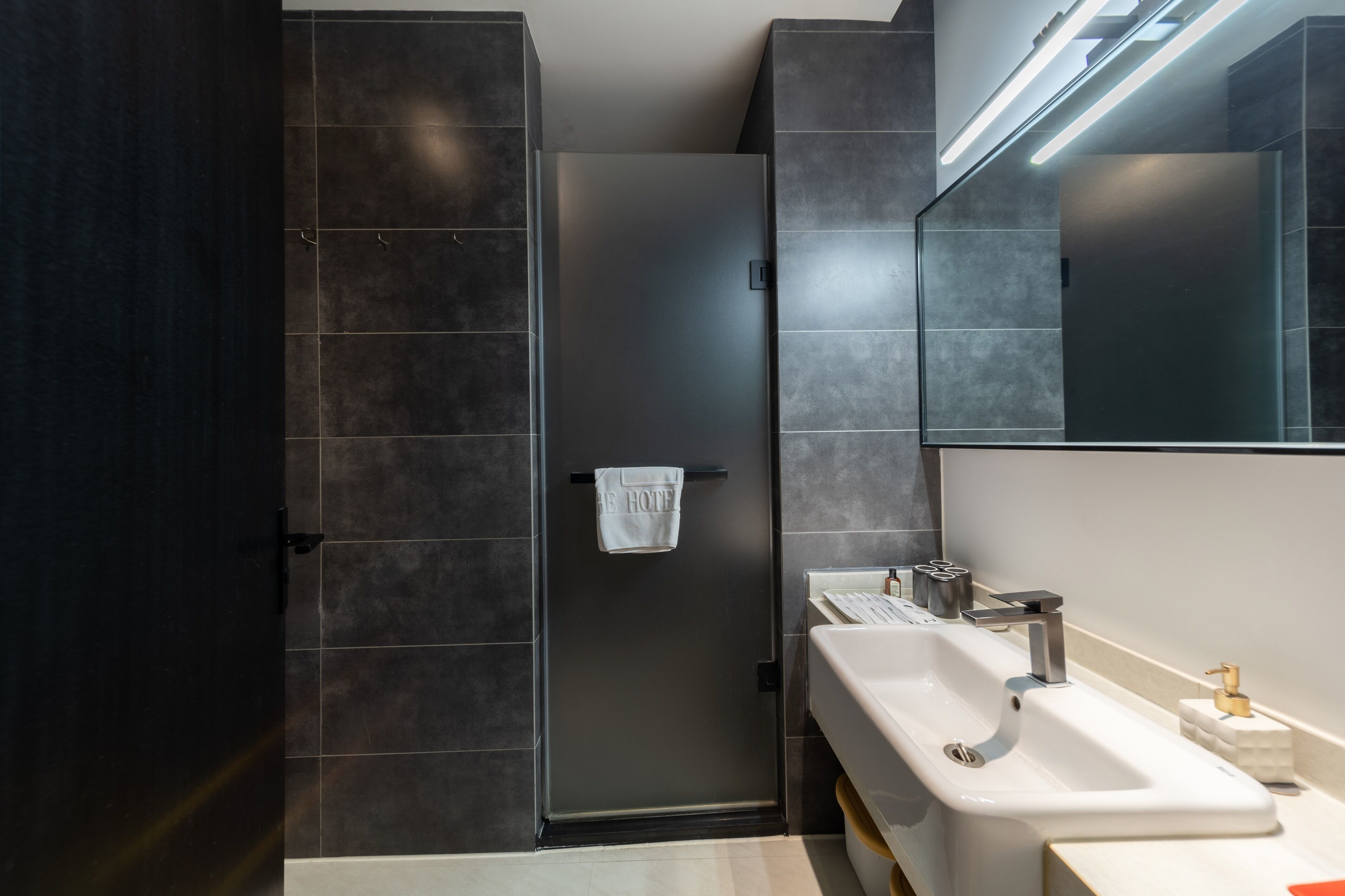 A modern bathroom with dark tiles, a white sink, and a towel hanging on a door.