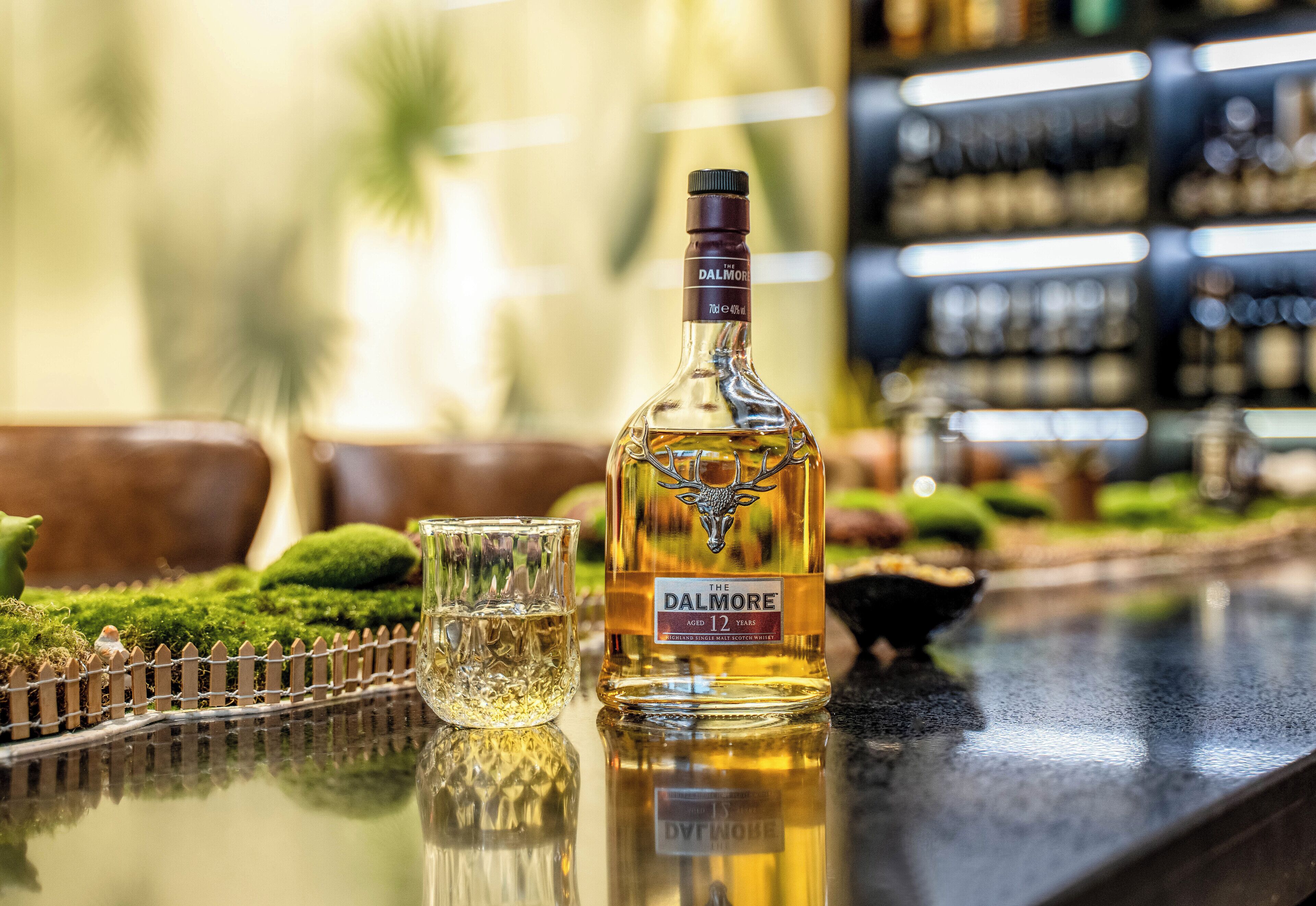 A bottle of Dalmore 12 years old whiskey on a bar counter with a glass and a decorative fence in the background.