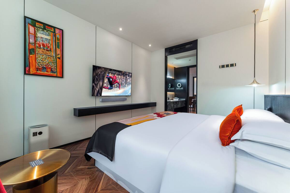 Une chambre d’hôtel moderne dotée d’un grand lit, d’un téléviseur à écran plat fixé au mur, d’une petite table d’appoint et d’une œuvre d’art originale au-dessus du lit.