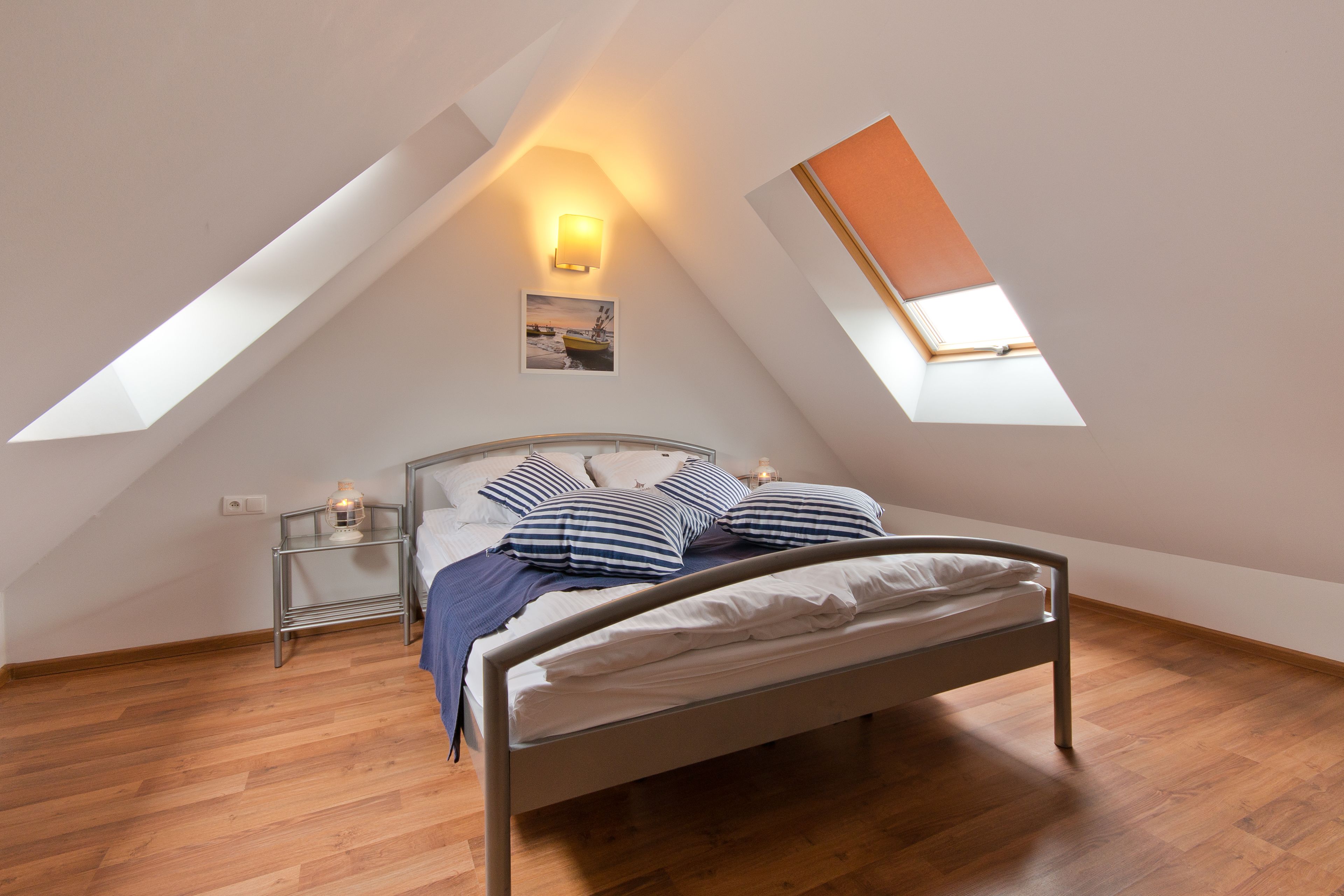 A bedroom with a sloped ceiling, a bed with white bedding, striped pillows, a nightstand with a lamp, and a framed picture on the wall.