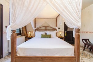 Una cama con dosel, ropa de cama blanca y una almohada verde, un cabecero de madera y una mesita de noche con una lámpara.
