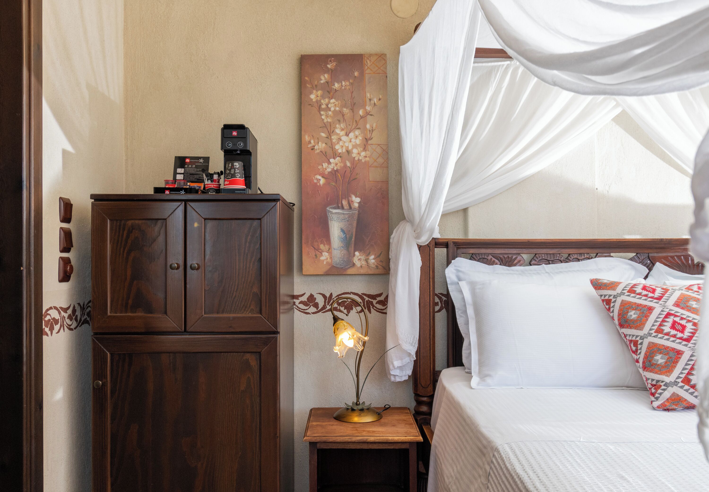 A bedroom with a wooden cabinet, a bedside table with a lamp, a bed with white bedding and patterned pillows, and a painting of flowers on the wall.