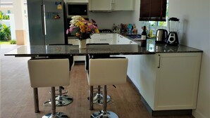 A kitchen with a granite countertop, white cabinets, a refrigerator, a microwave, a sink, a coffee maker, and a blender. There are two bar stools and a vase with flowers on the countertop. - Villa de Veluwe at Hua Hin (Hua Hin)