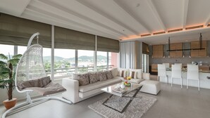 Uma sala de estar moderna com um sofá branco, uma cadeira suspensa, uma mesa de centro e vista para o horizonte urbano através de amplas janelas.