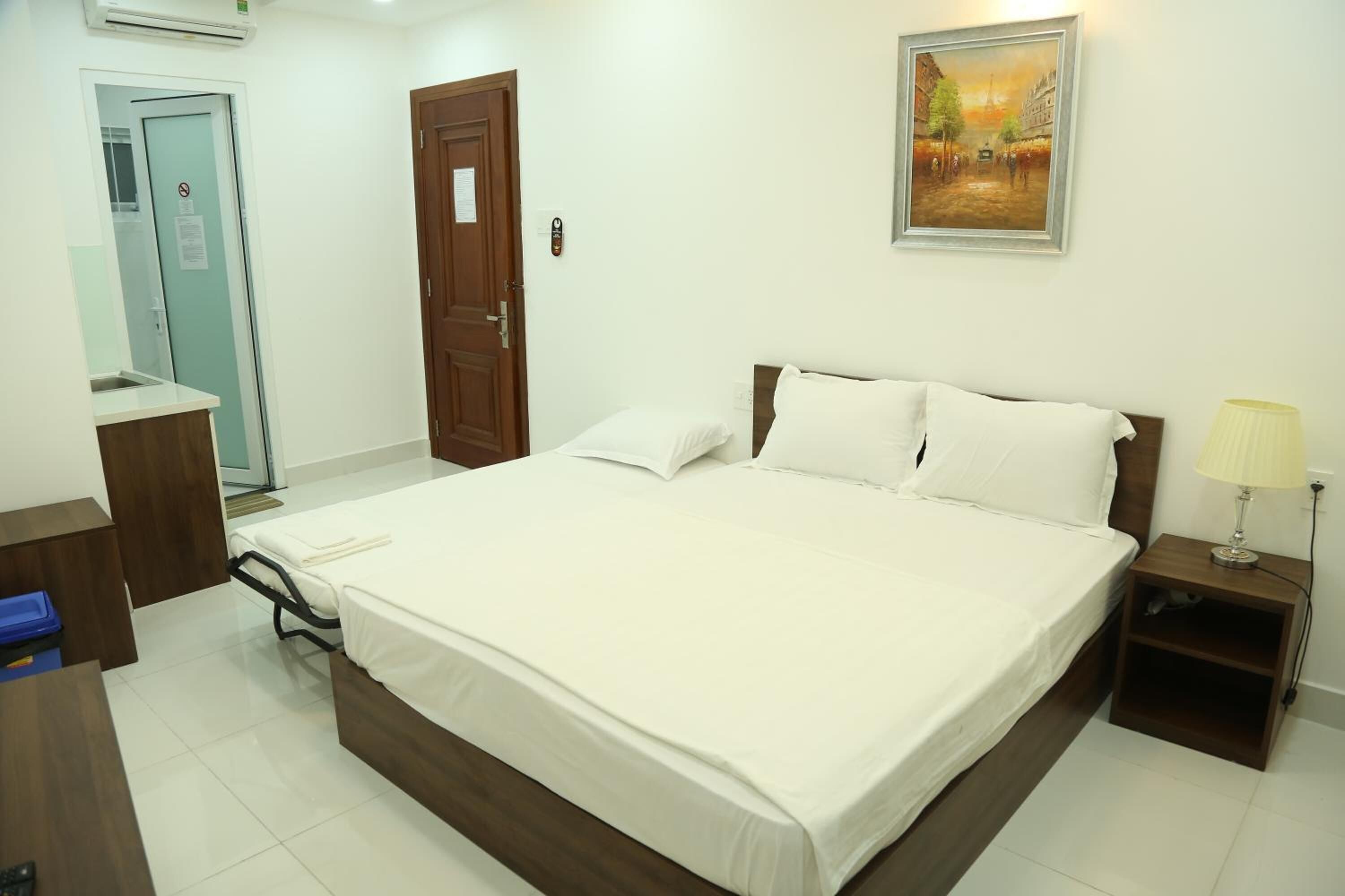 A hotel room with a large bed, a desk, a chair, a nightstand with a lamp, and a painting on the wall.