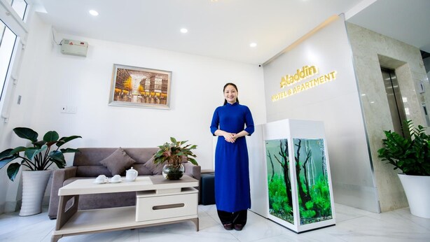 Un hall d’hôtel comprenant un comptoir de réception avec un réceptionniste, un canapé, une table basse, des plantes en pot, un aquarium et un lustre.