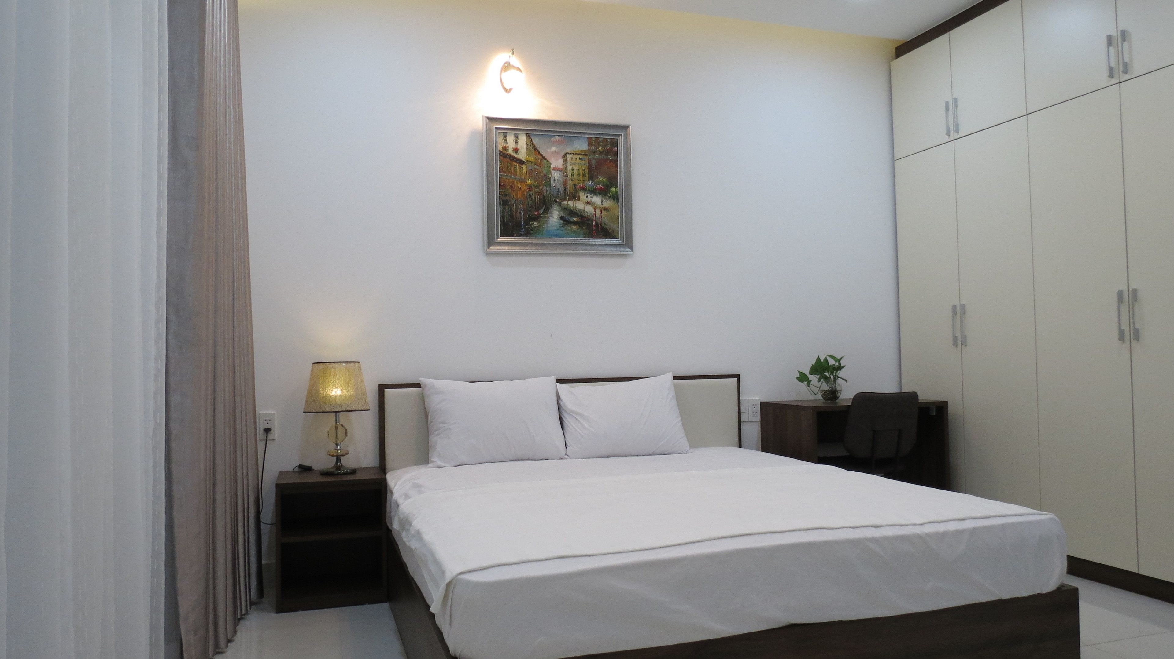 A hotel room with a bed, a nightstand with a lamp, a painting on the wall, a desk with a chair, and a wardrobe.