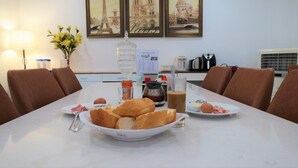 Une table à manger avec du pain, des assiettes et des condiments, dans une pièce où se trouvent des chaises marron, une lampe et des photos encadrées de monuments parisiens.