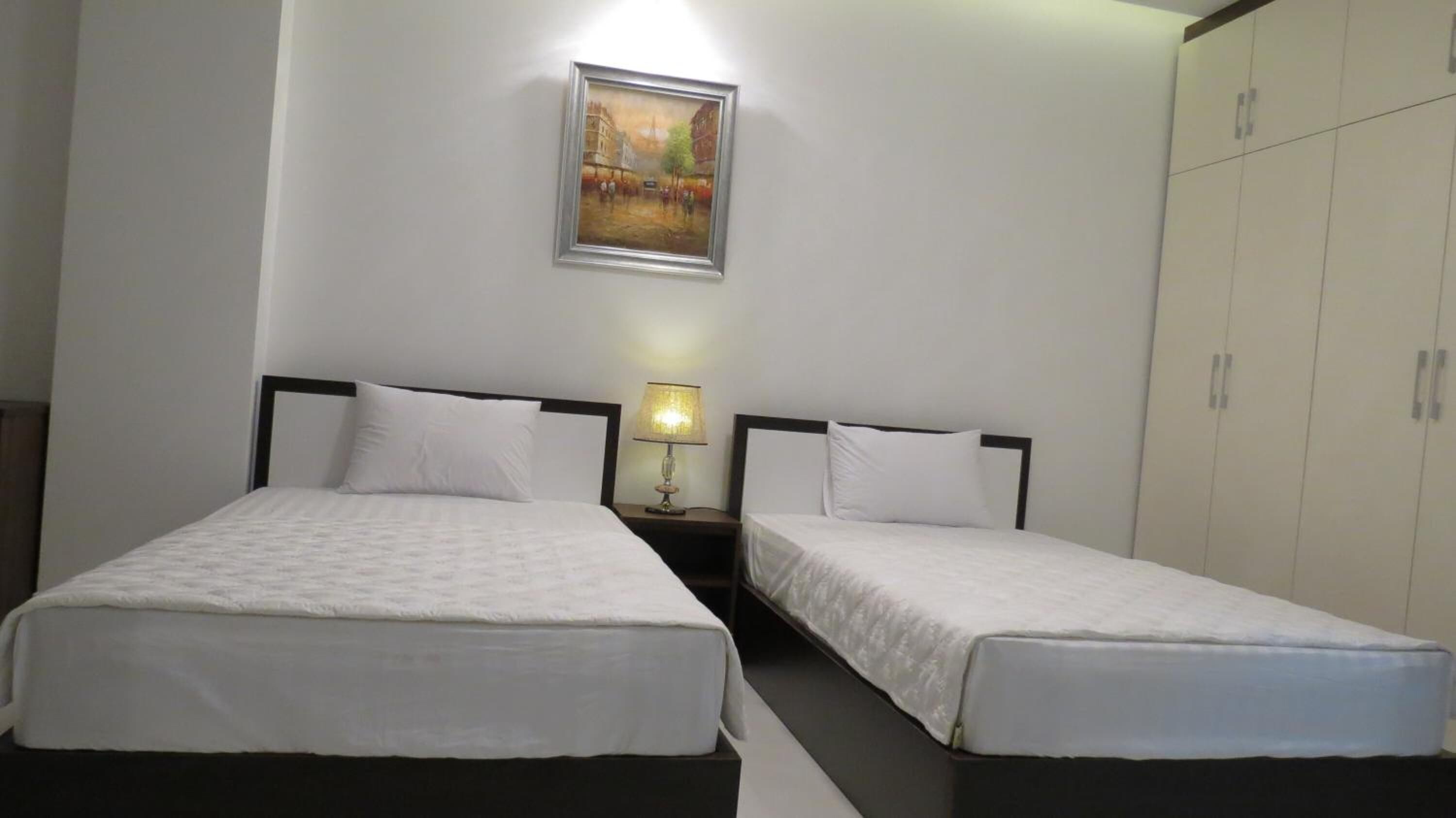 A hotel room with two single beds, a nightstand with a lamp, a painting on the wall, and a wardrobe.