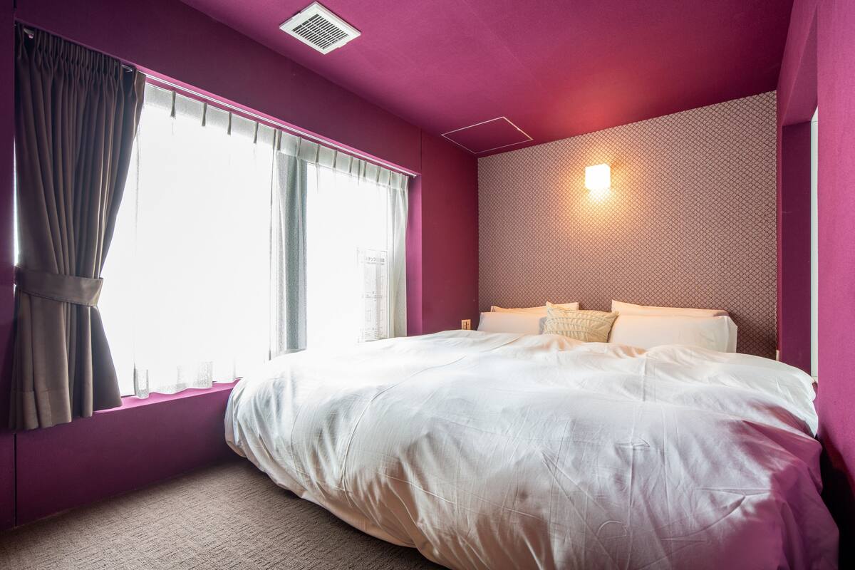 A bedroom with a large bed, a window with curtains, a wall-mounted light, and a patterned wall.
