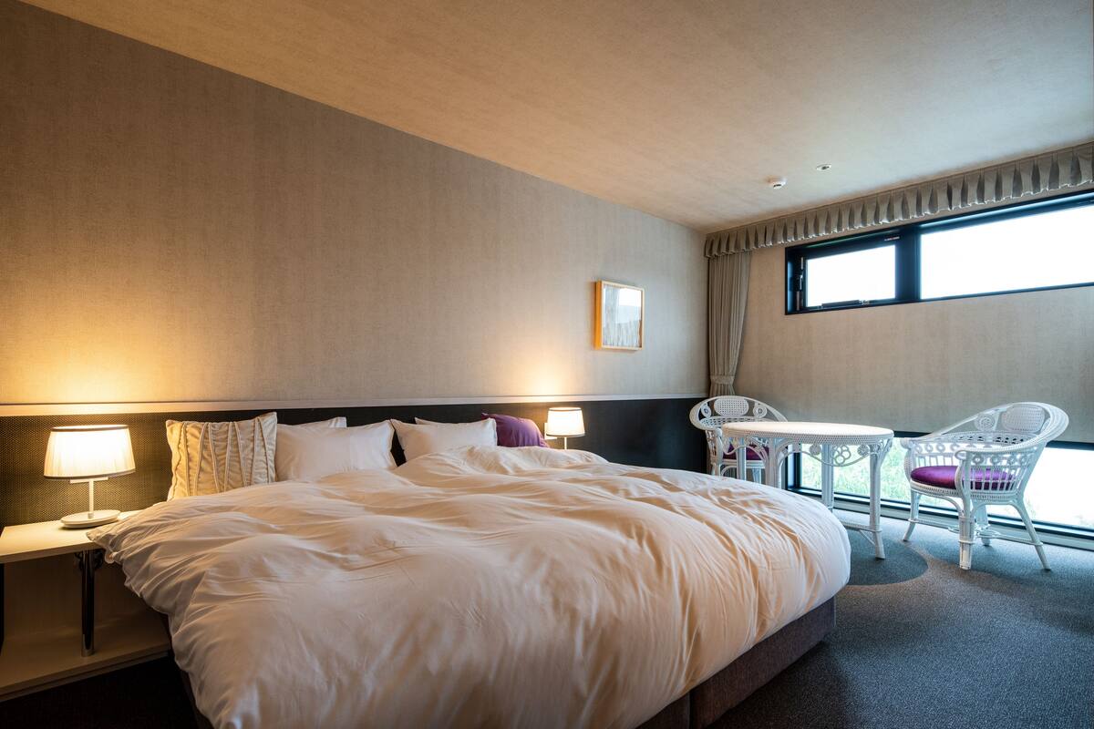 A hotel room with a large bed, a small table, a chair, and a window with curtains.