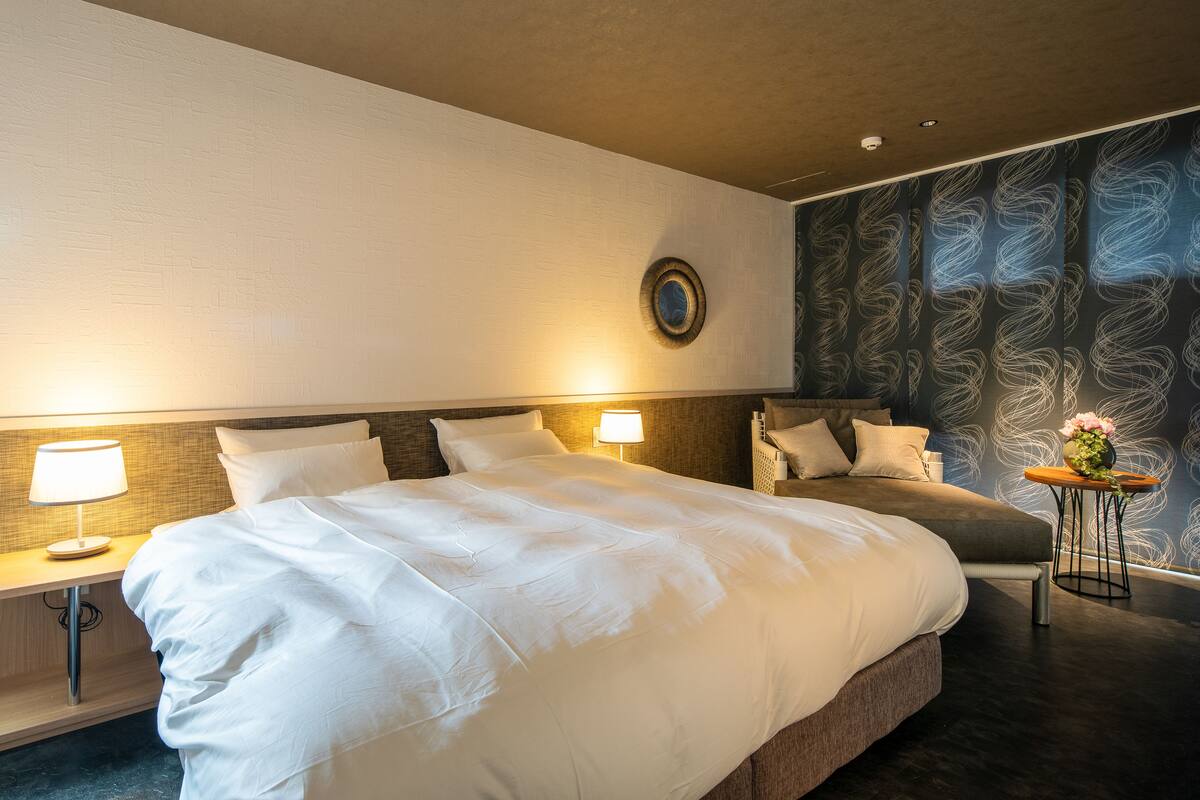 A hotel room with a large bed, two bedside tables with lamps, a small side table with a vase, and a patterned wall.