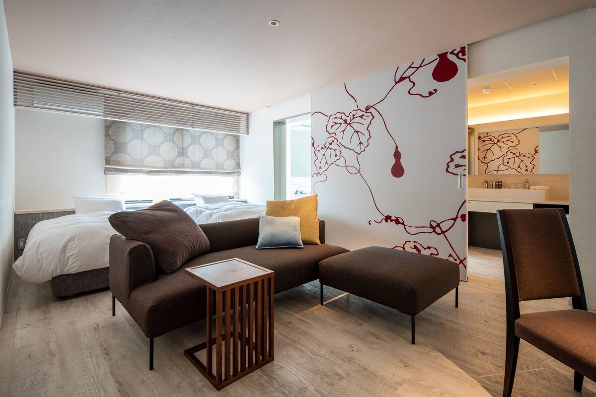 A modern hotel room with a large bed, a brown sofa, a wooden side table, and a large mirror.