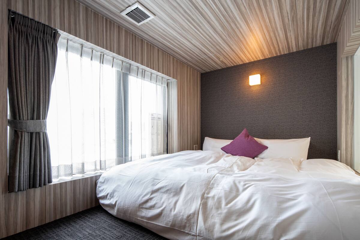A hotel room with a large bed, a window with curtains, a headboard with a light fixture, and a textured wall.