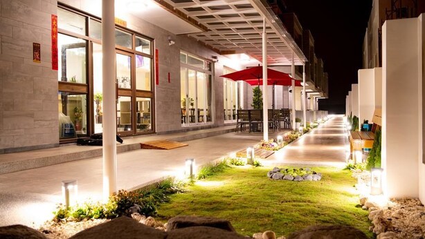 An outdoor area with a covered seating area, a pathway, and a building with large windows and a red umbrella. - Brother B&B (Magong)