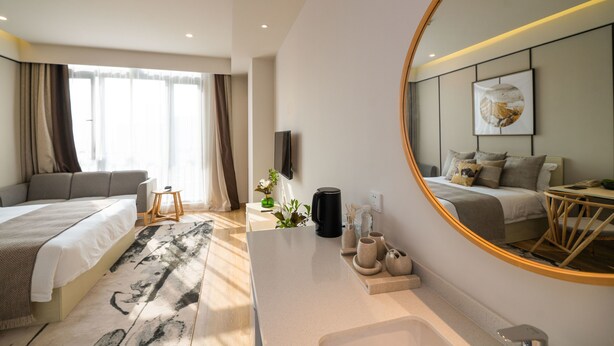 A modern hotel room with a large bed, a sitting area, a small table, a coffee maker, and a mirror reflecting the room. - Suisse Place Shanghai (Shanghai)