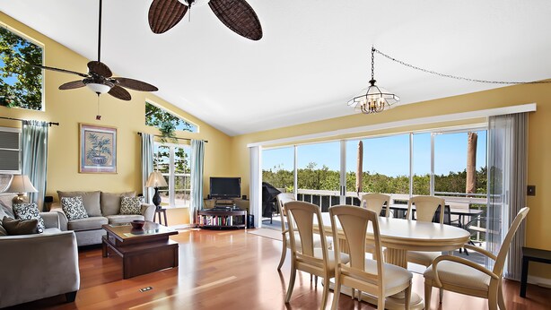 A spacious living room with a large dining table, comfortable seating, a ceiling fan, and large windows offering a view of the outdoors. - Sherwood Lakes A (Palm Harbor)