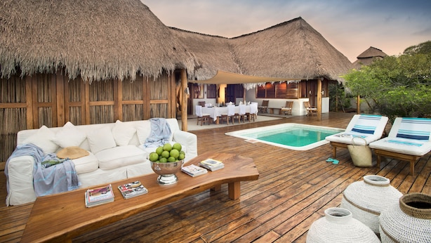 Une terrasse en bois avec une piscine, des chaises longues blanches et un canapé blanc, le tout agrémenté d’une structure de toit en chaume en arrière-plan.