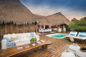 A wooden deck with a pool, white lounge chairs, and a white sofa with a thatched roof structure in the background.
