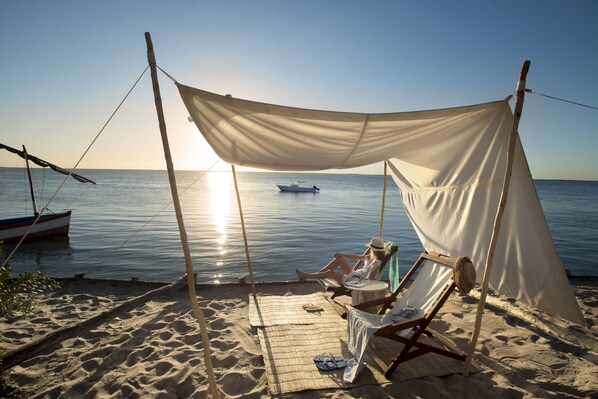A beach with a canopy, a boat, and a person relaxing on a chair.