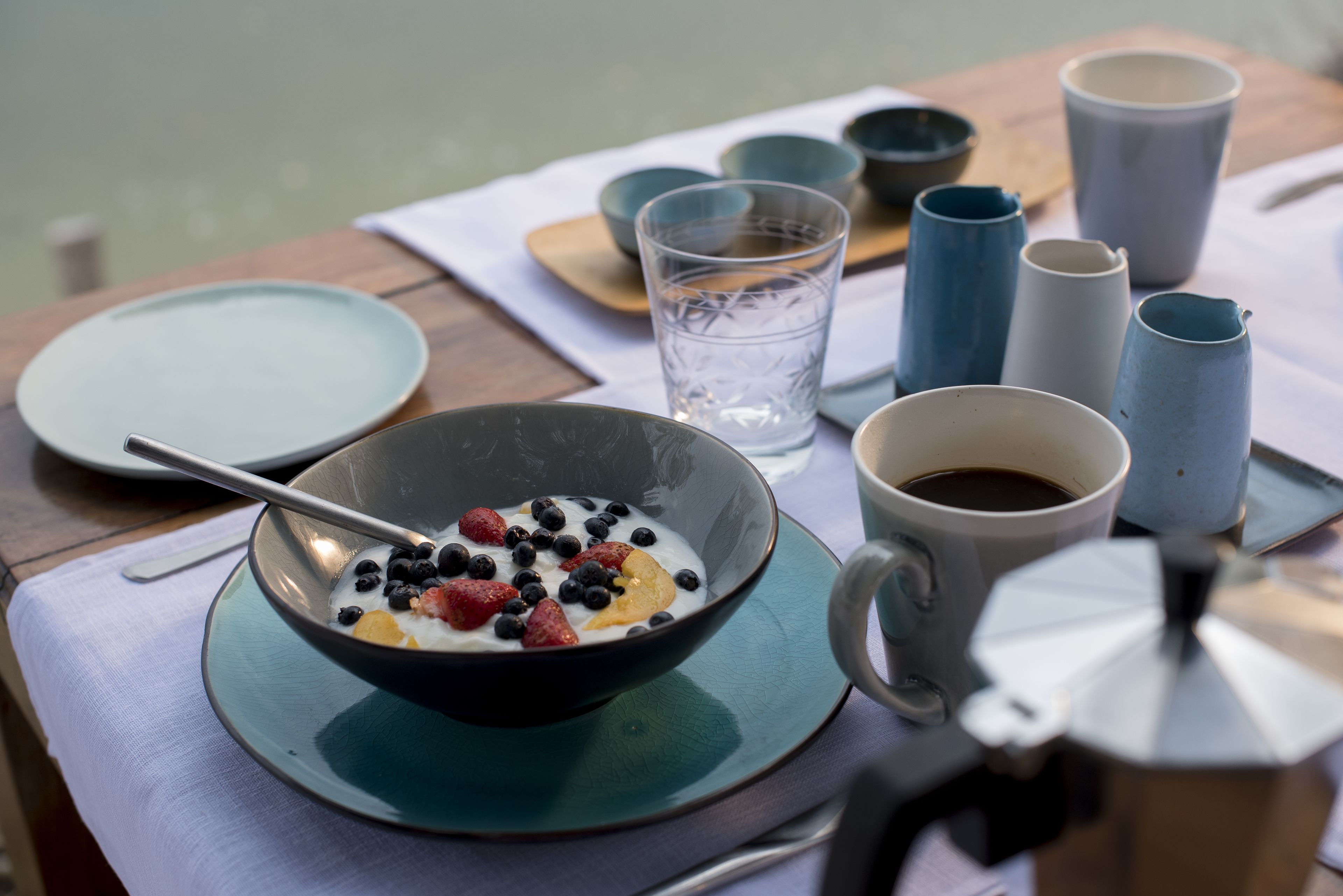 Desayuno con un tazón de yogur cubierto de bayas, una taza de café y varias tazas y recipientes sobre una mesa.
