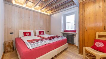 A bedroom with a wooden ceiling, a bed with red bedding, a wooden chair, and a window with a view of the outside.