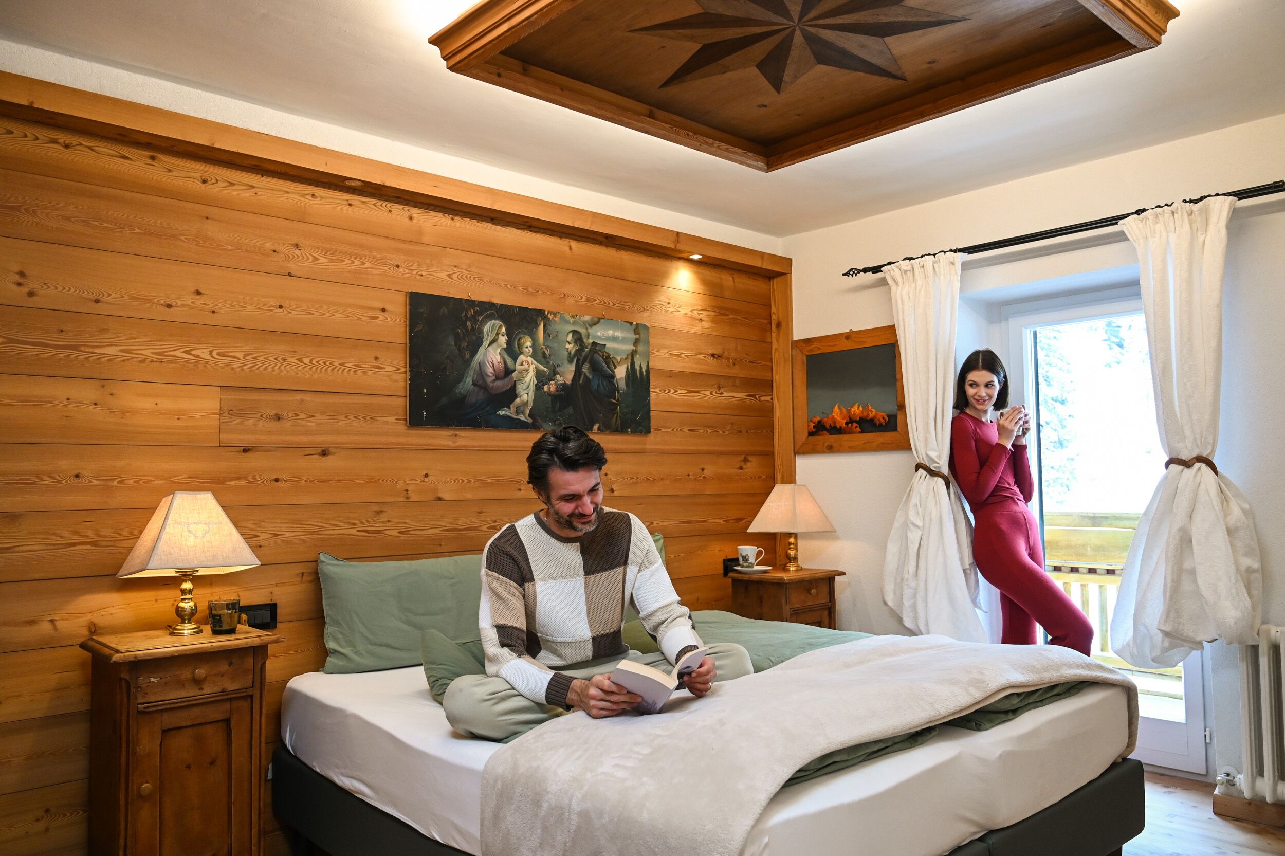 A bedroom with a wooden wall, a bed with a green bedspread, two bedside tables with lamps, and a window with curtains.