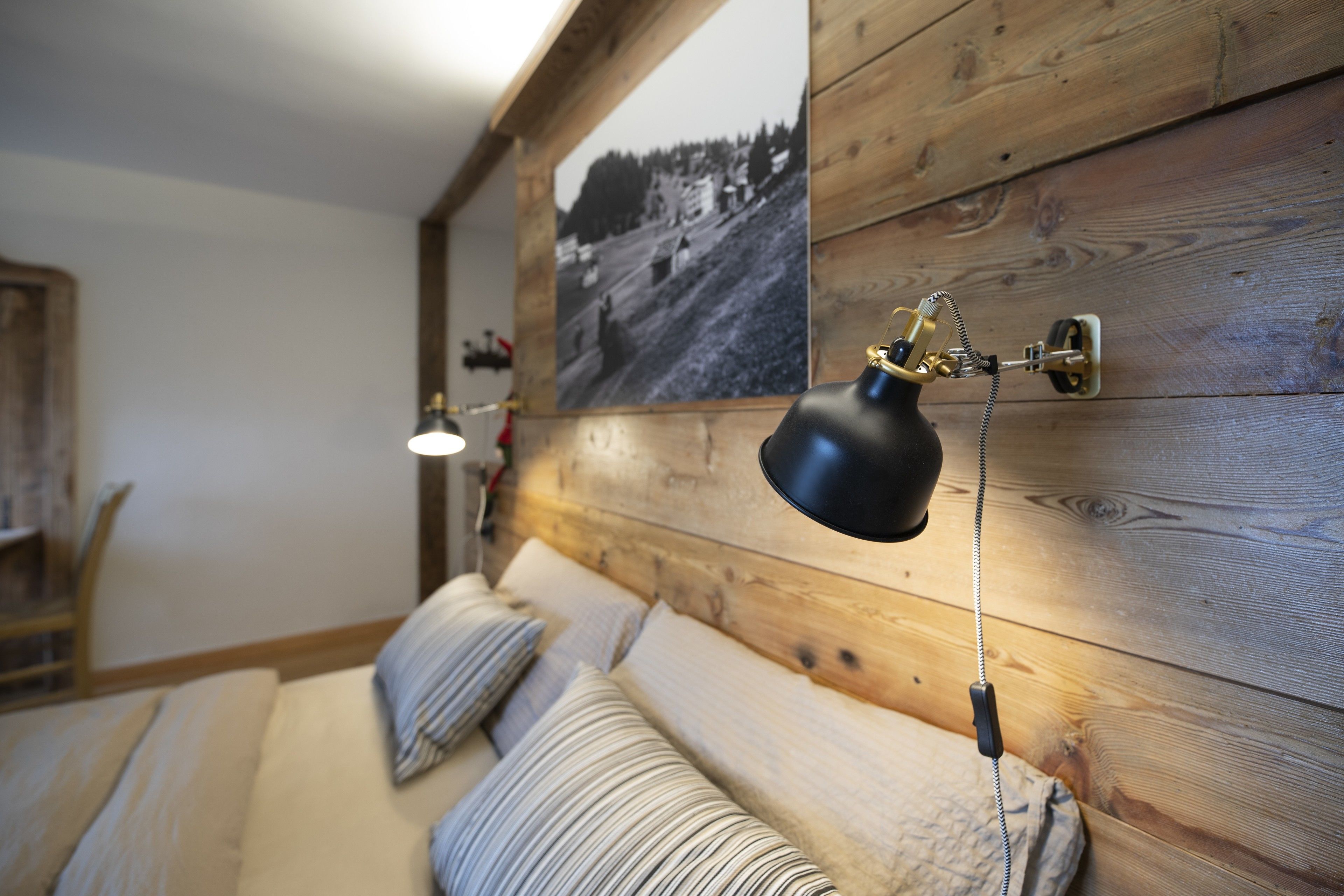 Une chambre chaleureuse avec une tête de lit en bois, un lit agrémenté d’oreillers à rayures et une lampe fixée au mur.
