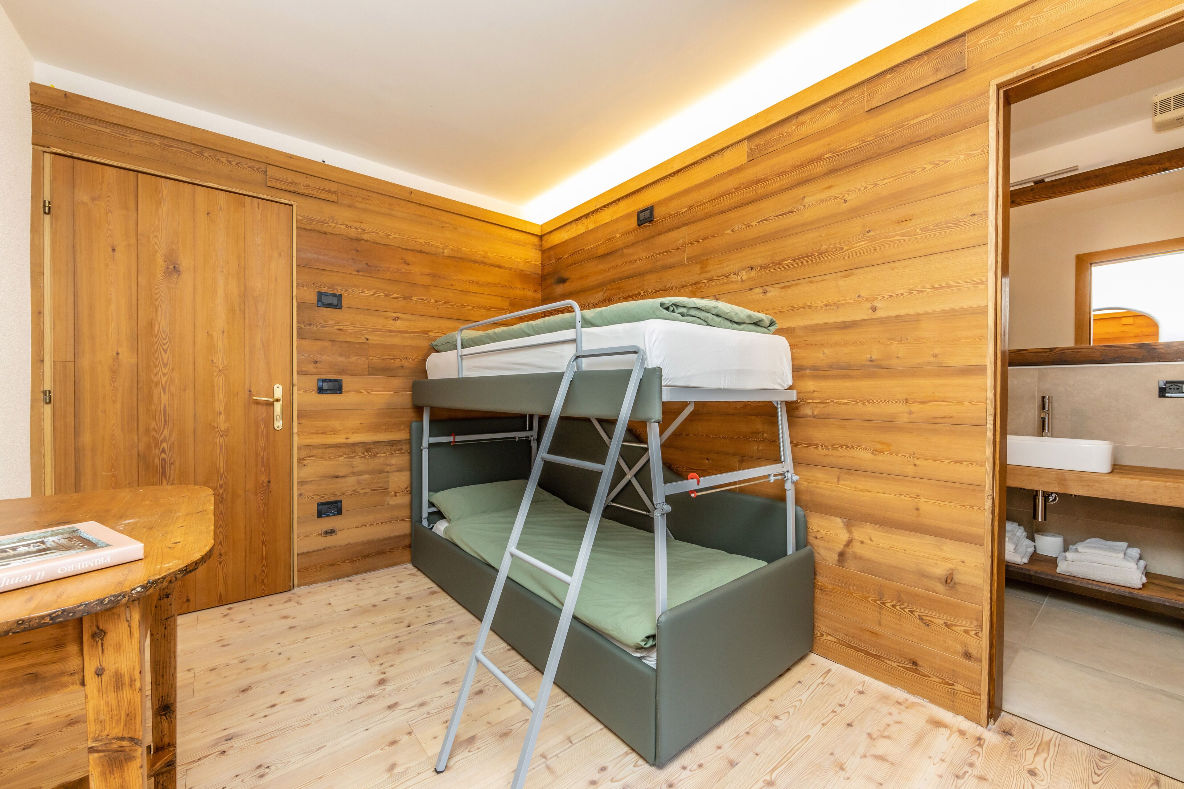 A bunk bed with a ladder, wooden walls, a wooden door, a small table with a book, and a bathroom with a sink and towel rack.