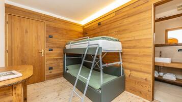 A bunk bed with a ladder, wooden walls, a wooden door, a small table with a book, and a bathroom with a sink and towel rack.