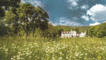 A large, white, multi-story house with a symmetrical facade, surrounded by a lush green field with tall grass and wildflowers, under a partly cloudy sky.