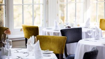 Un comedor con mesas dispuestas para dos, con manteles blancos y cubertería ordenada.