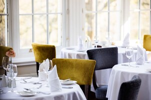 Un comedor con mesas dispuestas para dos, con manteles blancos y cubertería ordenada.