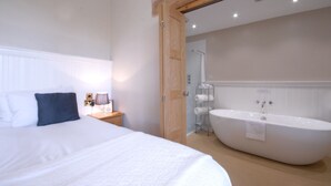 Een slaapkamer met een groot bed, een nachtkastje met een lamp en een badkamer met een vrijstaand bad.