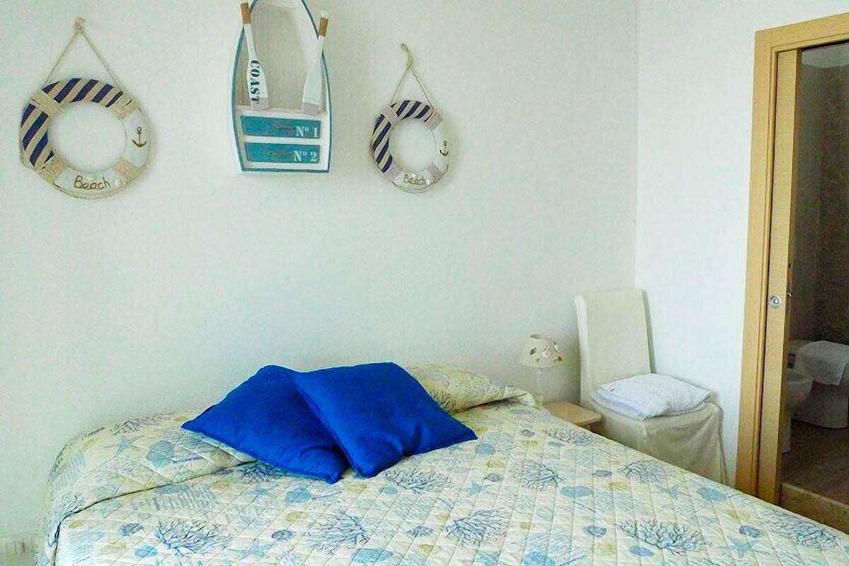 Un dormitorio con cama, mesita de noche, una silla y una puerta que da a un baño.