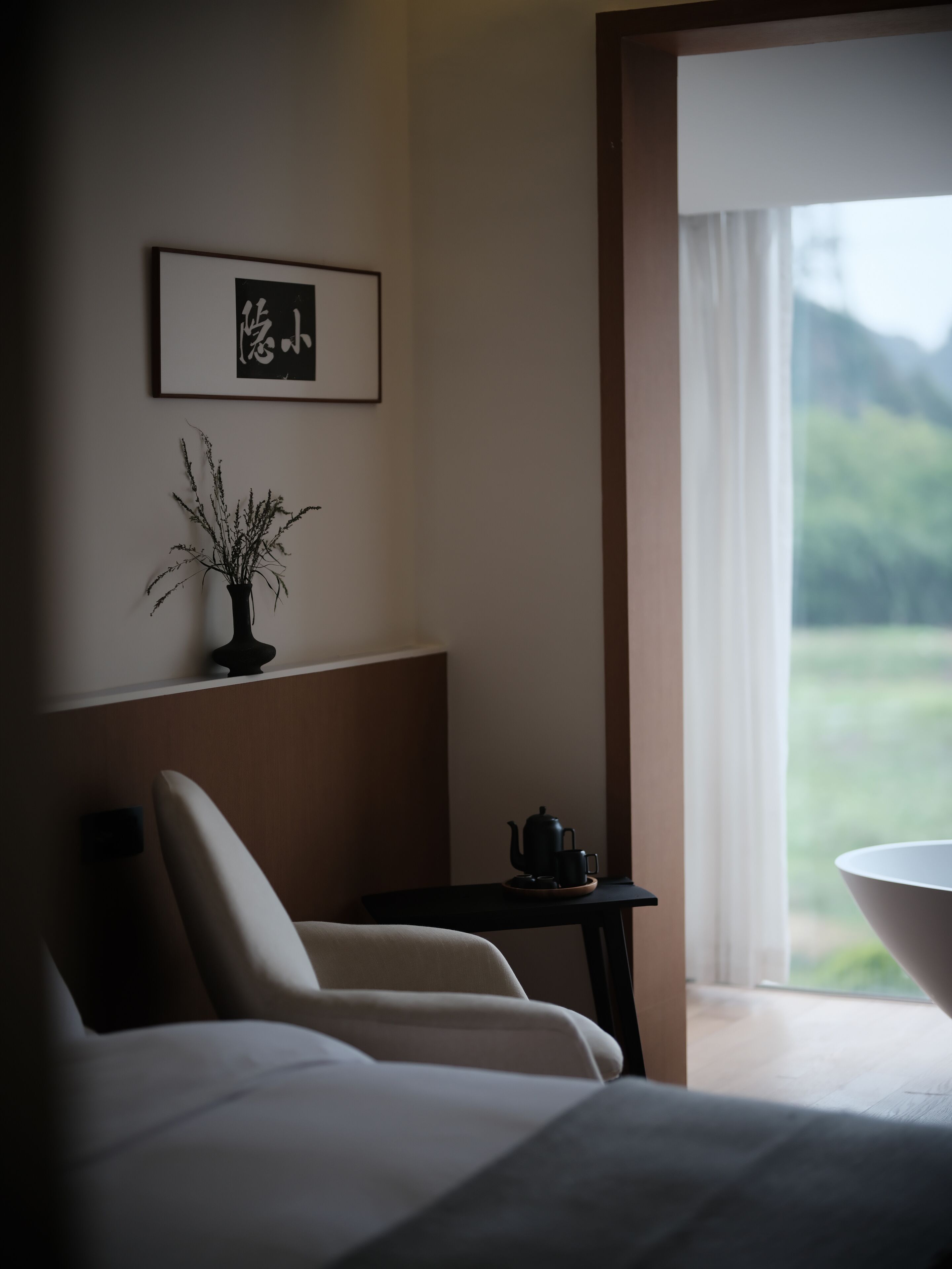 A hotel room with a bed, a small table with a teapot and cup, a framed picture on the wall, and a view of greenery outside.