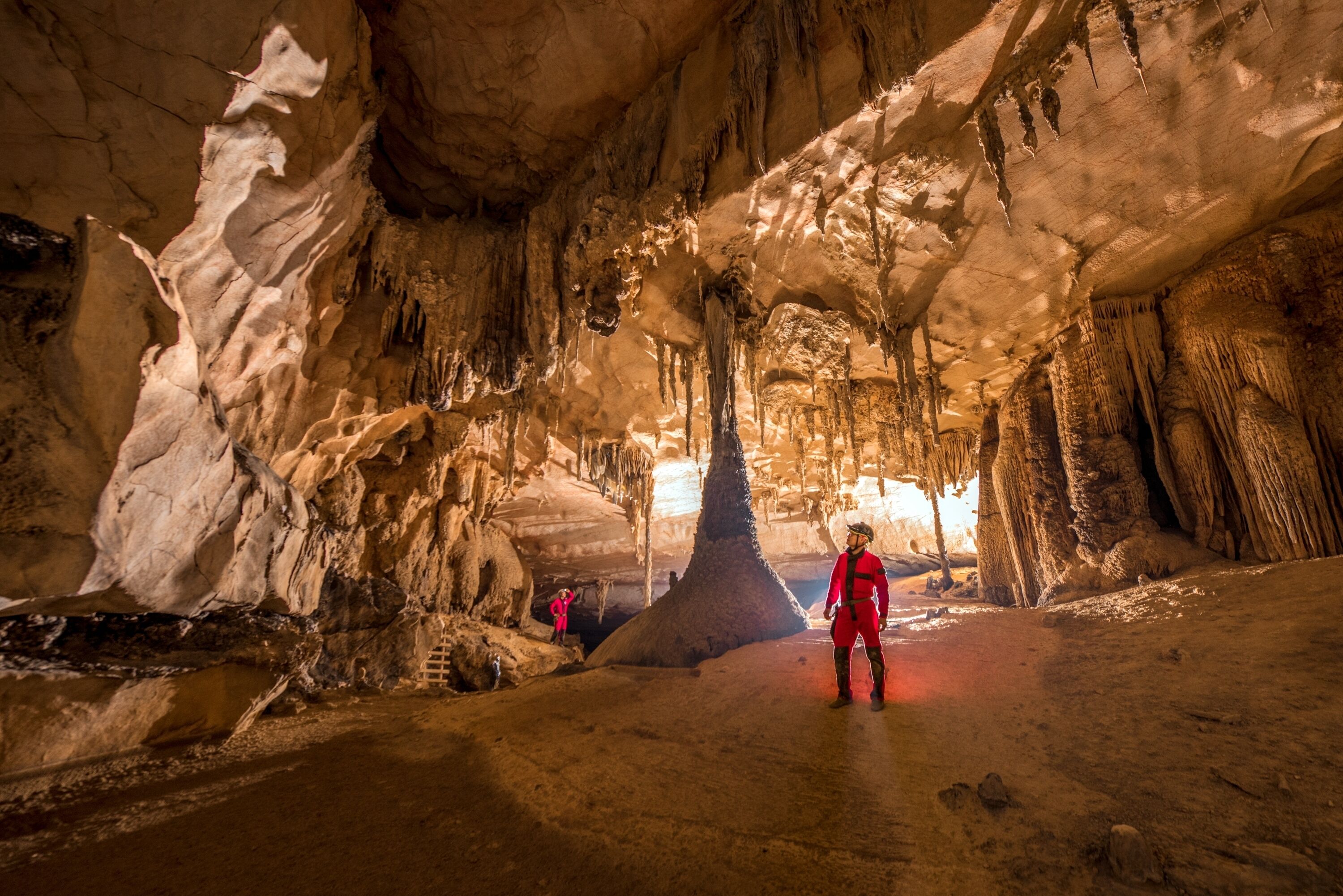 A cave interior with stalactites and stalagmites, a person in red, and another person in pink.